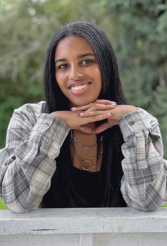 A smiling young woman with long straight hair, wearing a plaid shirt and layered necklaces, poses with her hands clasped in front of her against a blurred green background.