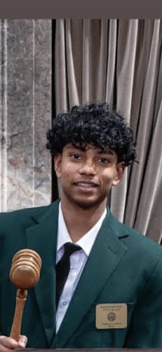 A young individual dressed in a green blazer, white shirt, and black tie, holding a gavel in front of a curtain backdrop.